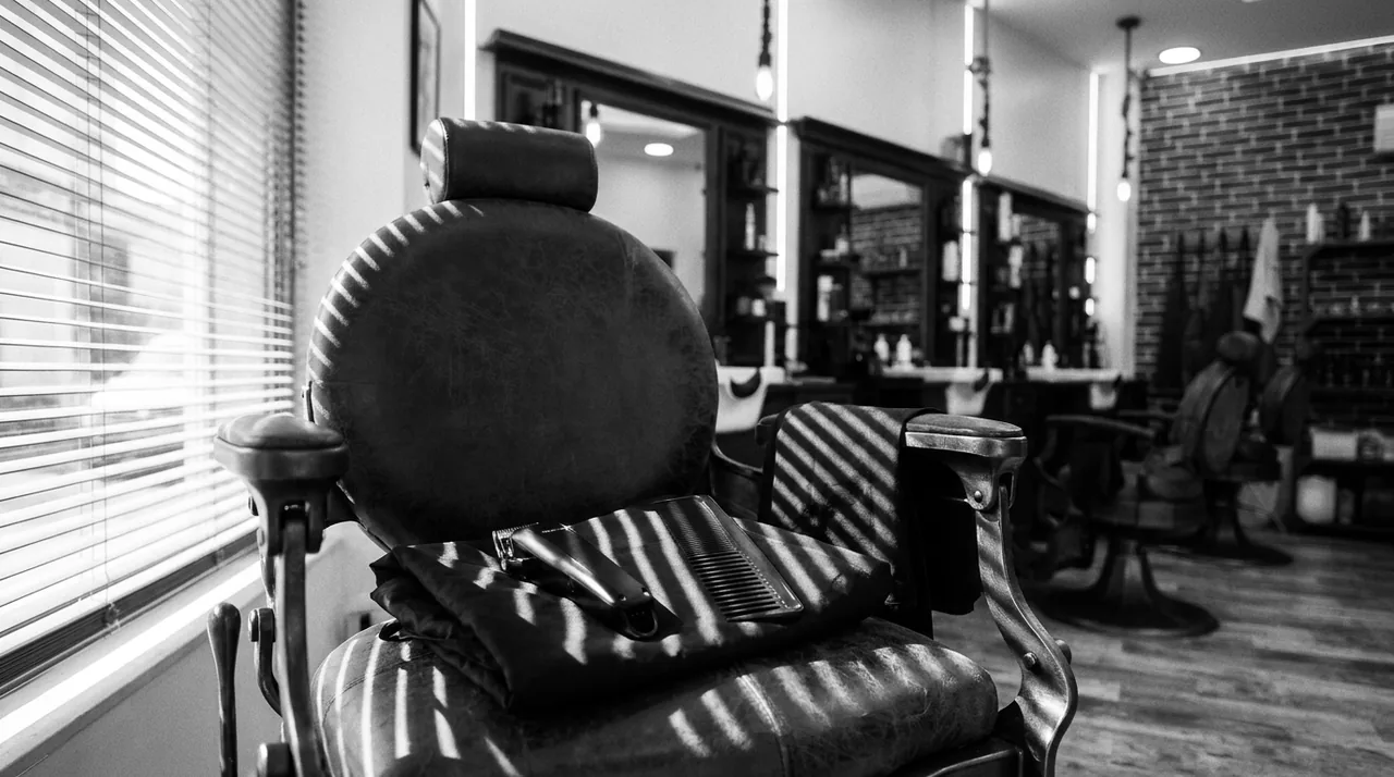 Cape noire pliée, tondeuse et peigne posés sur le siège d'un fauteuil de barbier vide, rayé par la lumière d'un store