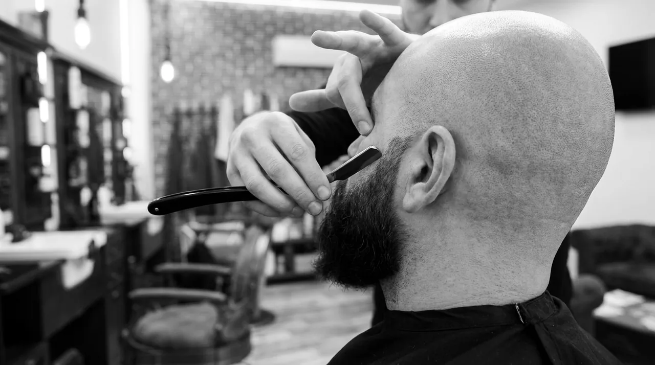 Vue par-dessus l'épaule : un rasoir trace la ligne haute d'une barbe foncée et épaisse