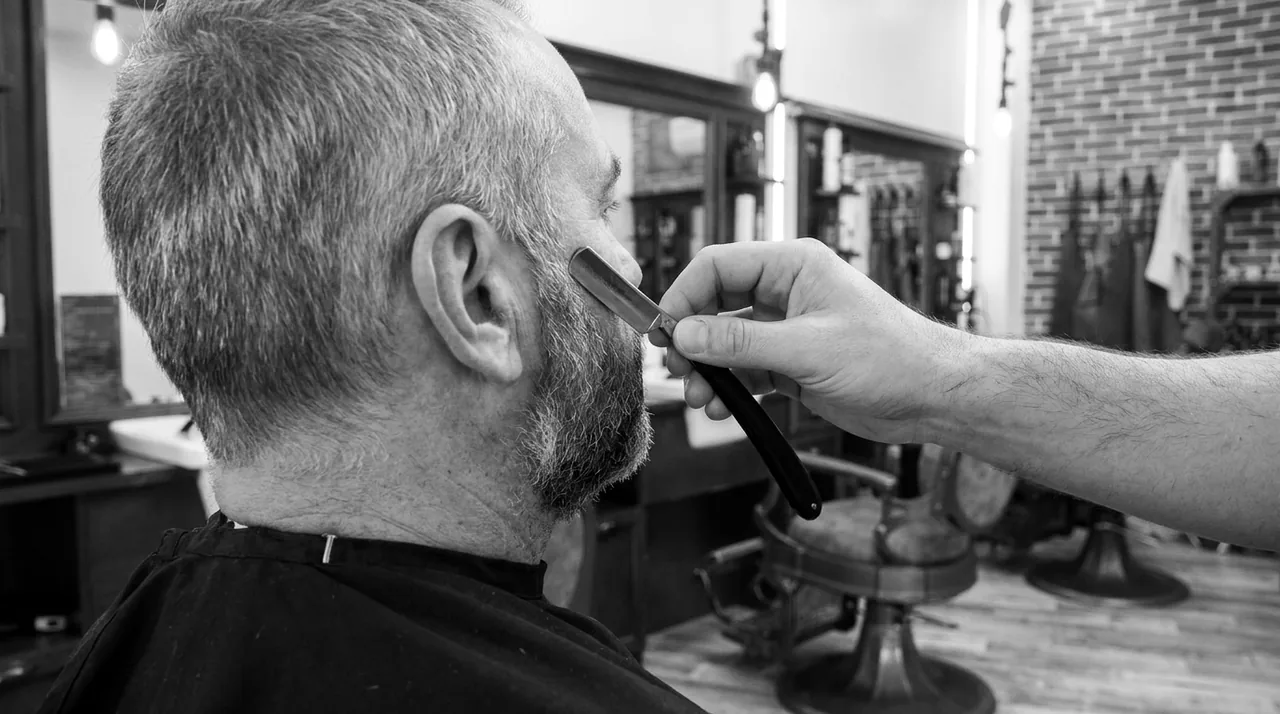 Vue par-dessus l'épaule : un rasoir trace le bord supérieur d'une barbe poivre et sel