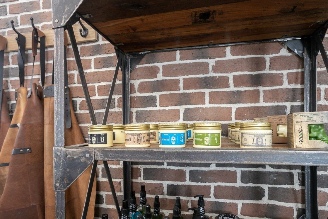 Pots de pommade coiffante dorés sur une étagère industrielle et tabliers en cuir devant un mur de briques chez Lui Coiffeur Draguignan