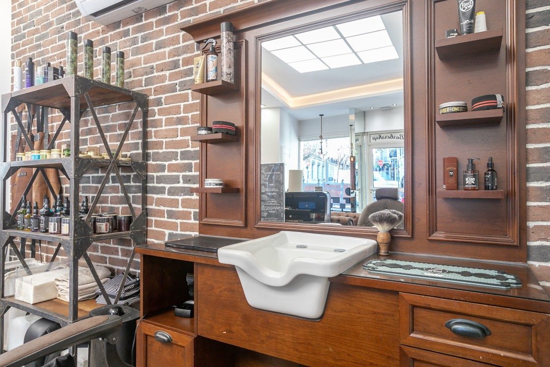 Poste de barbier en bois et bac blanc, miroir reflétant la vitrine sur rue, étagère de produits chez Lui Coiffeur Draguignan